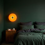 Orange acrylic donut lamp hung on a dark green bedroom wall above a bedside table at night