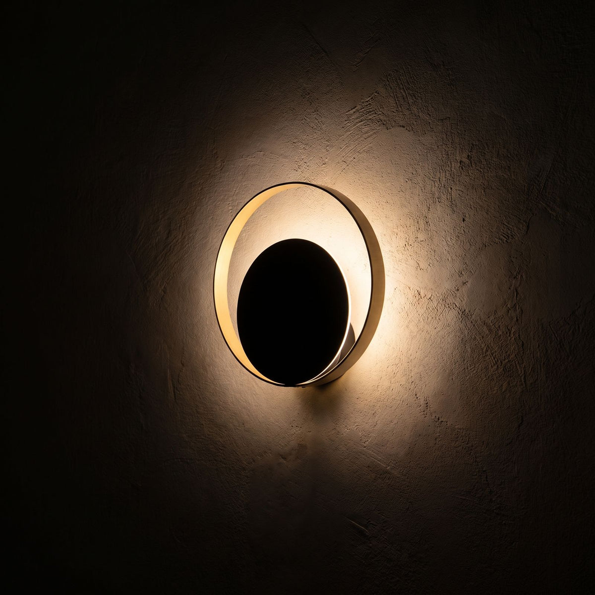 Brass Ring and Black Disc Wall Light glowing on a plaster wall at night