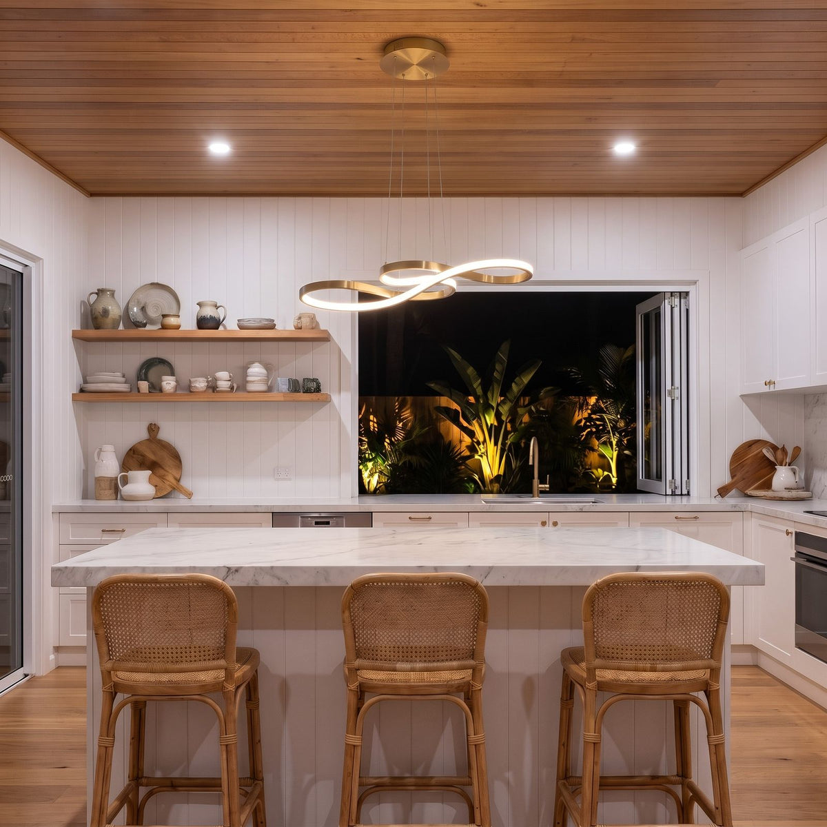 Gold Curved Ribbon Aluminium LED Pendant Light above a marble island bench with rattan stools in a kitchen at night