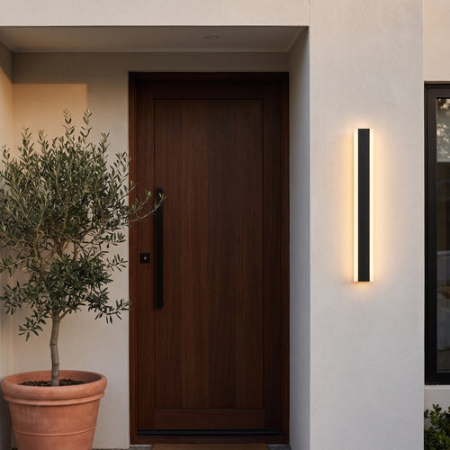 Linear LED wall light glowing warm white beside a timber front door and potted olive tree