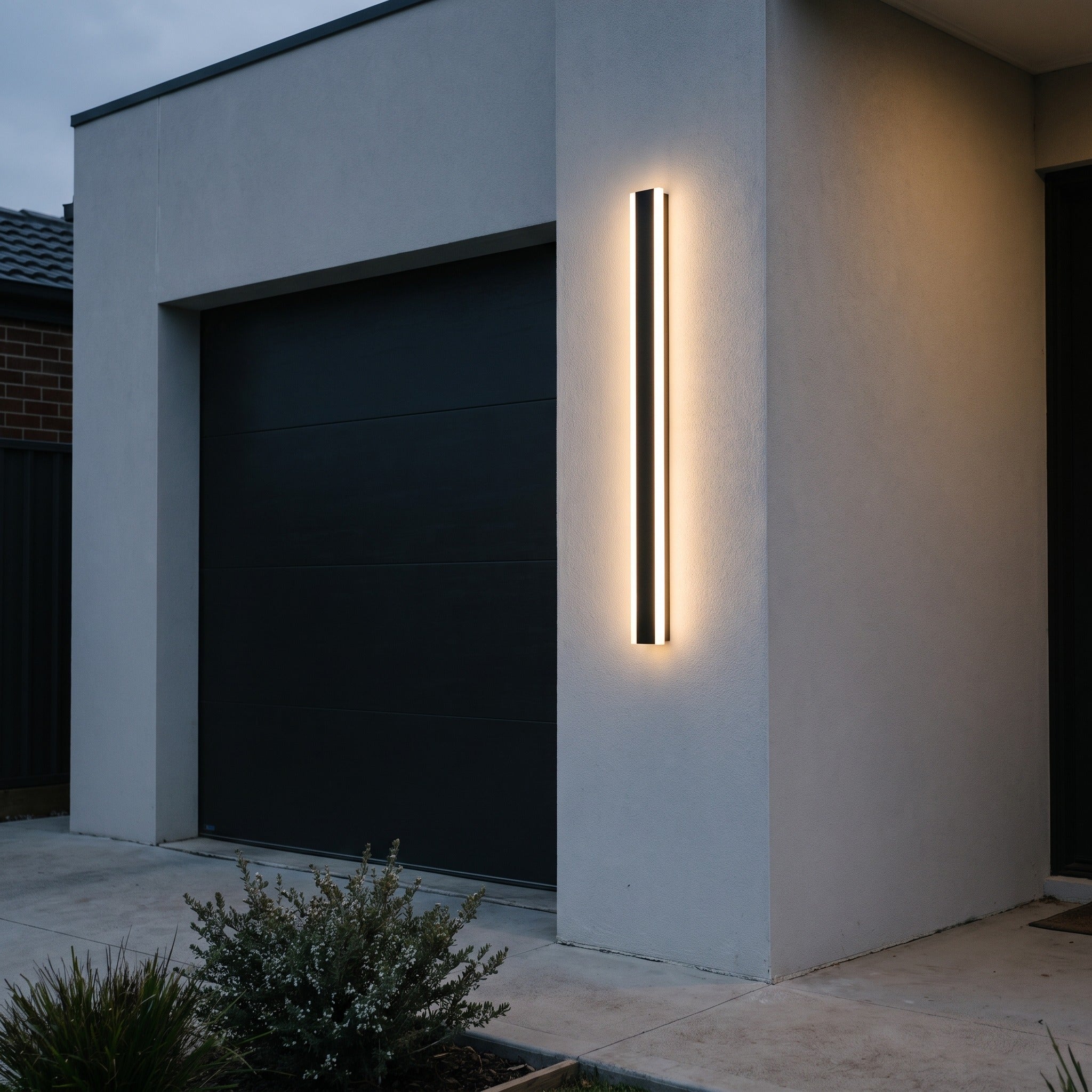 Linear LED wall light glowing on a grey rendered wall beside a dark garage door at dusk