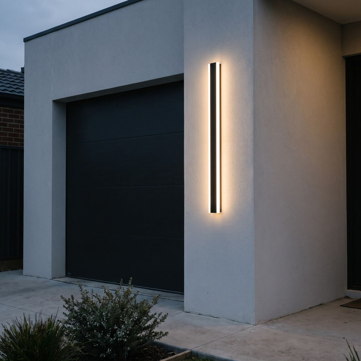 Linear LED wall light glowing on a grey rendered wall beside a dark garage door at dusk