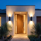 Two Flat Bar Aluminium Up Down Outdoor Wall Lights in black either side of a timber pivot door of a contemporary home at dusk