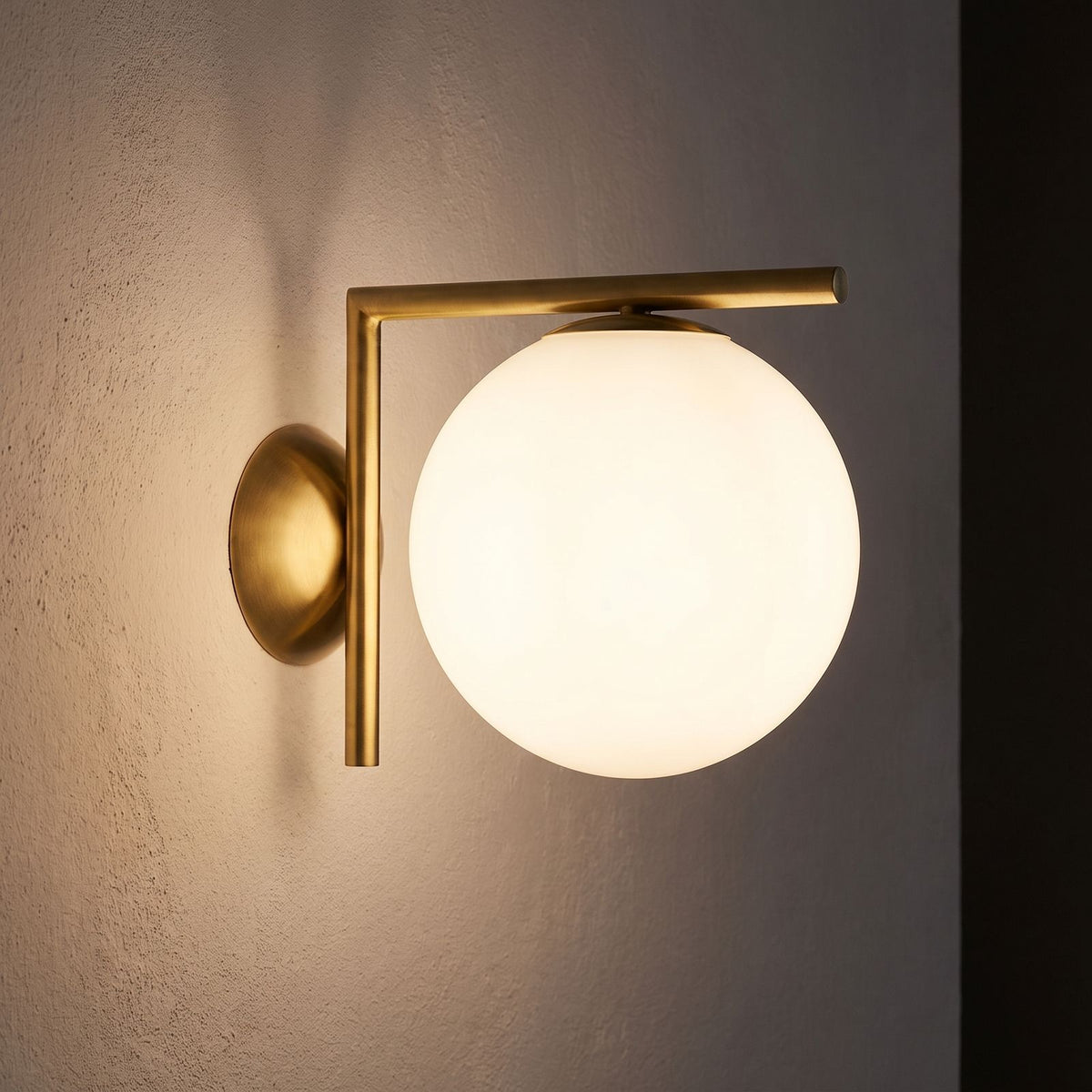 L-Arm Brass and White Glass Globe Wall Light glowing on a plaster wall at night