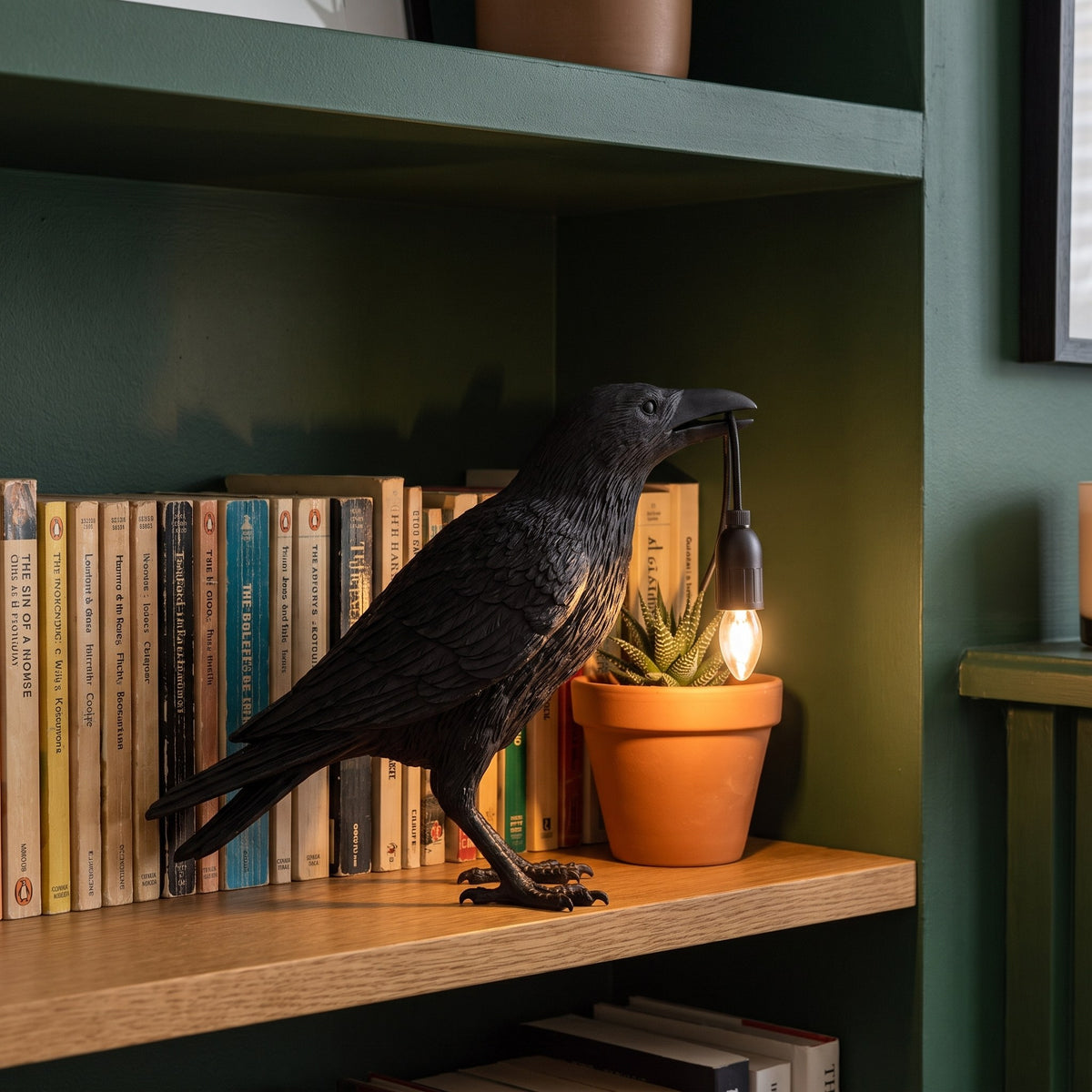 Raven Sculpture Resin Table Lamp lit on a bookshelf beside a row of books