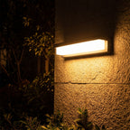 Rectangular Metal and Acrylic Outdoor Wall Light glowing on a garden wall at night