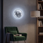 Round 3D Moon Black Frame LED Wall Light in cool white beside a velvet armchair in a reading nook at night