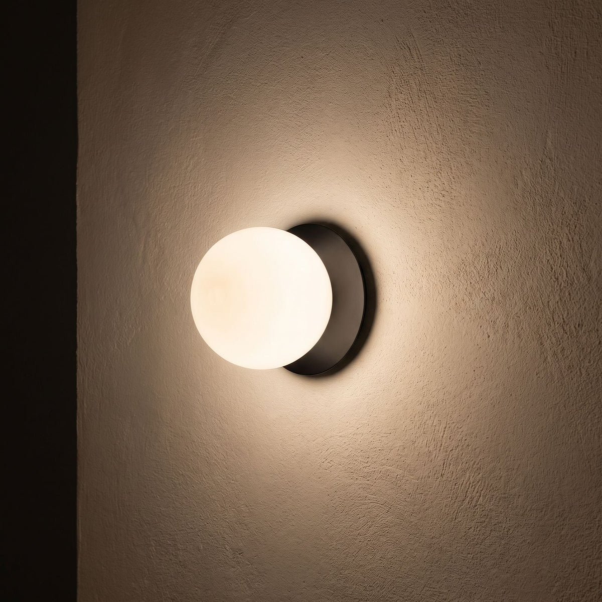 Round Glass Wall Sconce with Metal Backplate glowing on a plaster wall at night