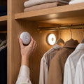 Hand placing a round rechargeable motion sensor wall light in a bedroom walk-in closet, a second one glowing warm white above the clothes