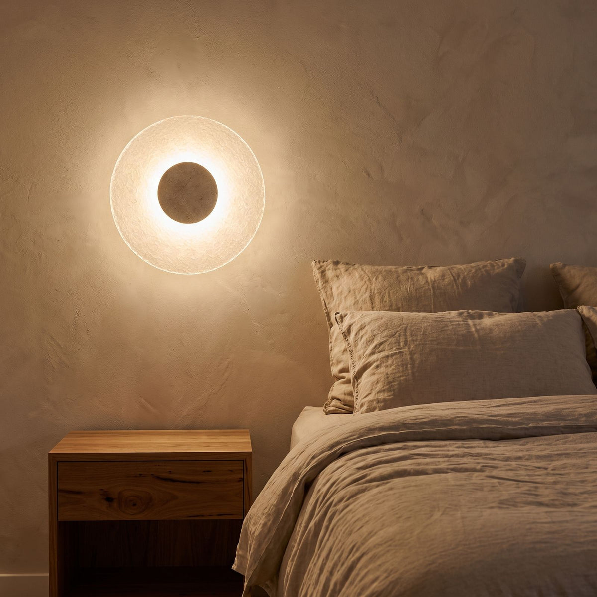 Round Travertine and Acrylic Wall Light beside a bed with linen bedding