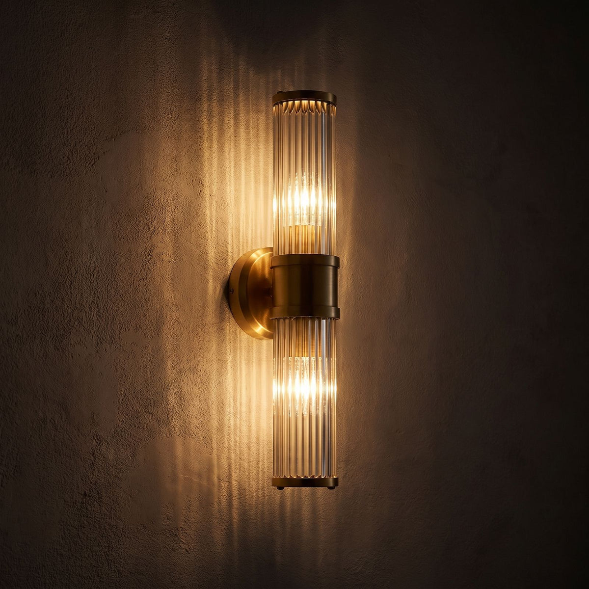 Ribbed Crystal Glass Tube Gold Wall Light with two tubes glowing on a plaster wall at night