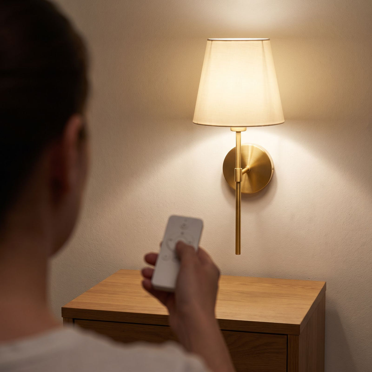 The wireless wall sconce in gold being dimmed with the included remote control beside a bed