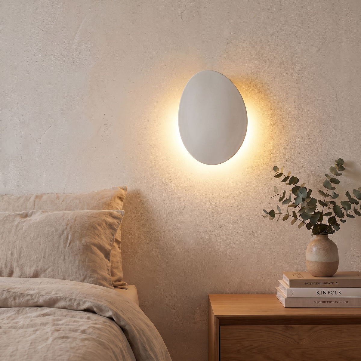 Tilted Pebble Aluminium Backlit LED Wall Light in white beside a bed