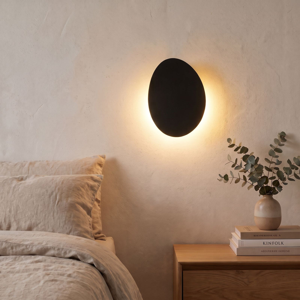 Tilted Pebble Aluminium Backlit LED Wall Light in black beside a bed
