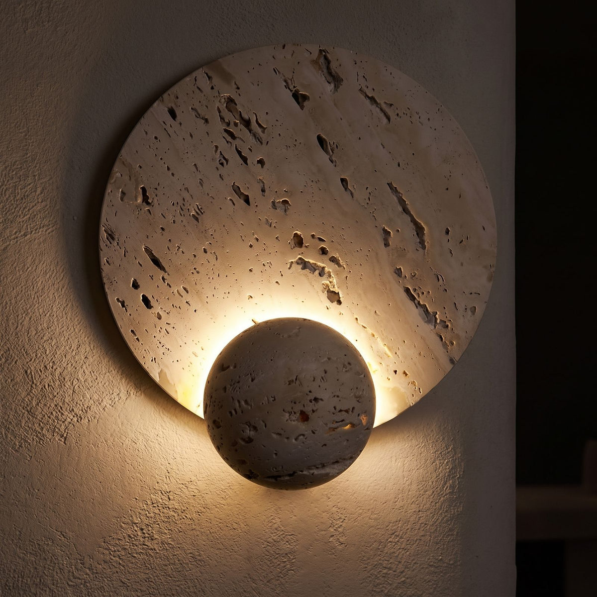 Travertine Pattern Marble Disc and Orb Wall Light glowing on a plaster wall at night