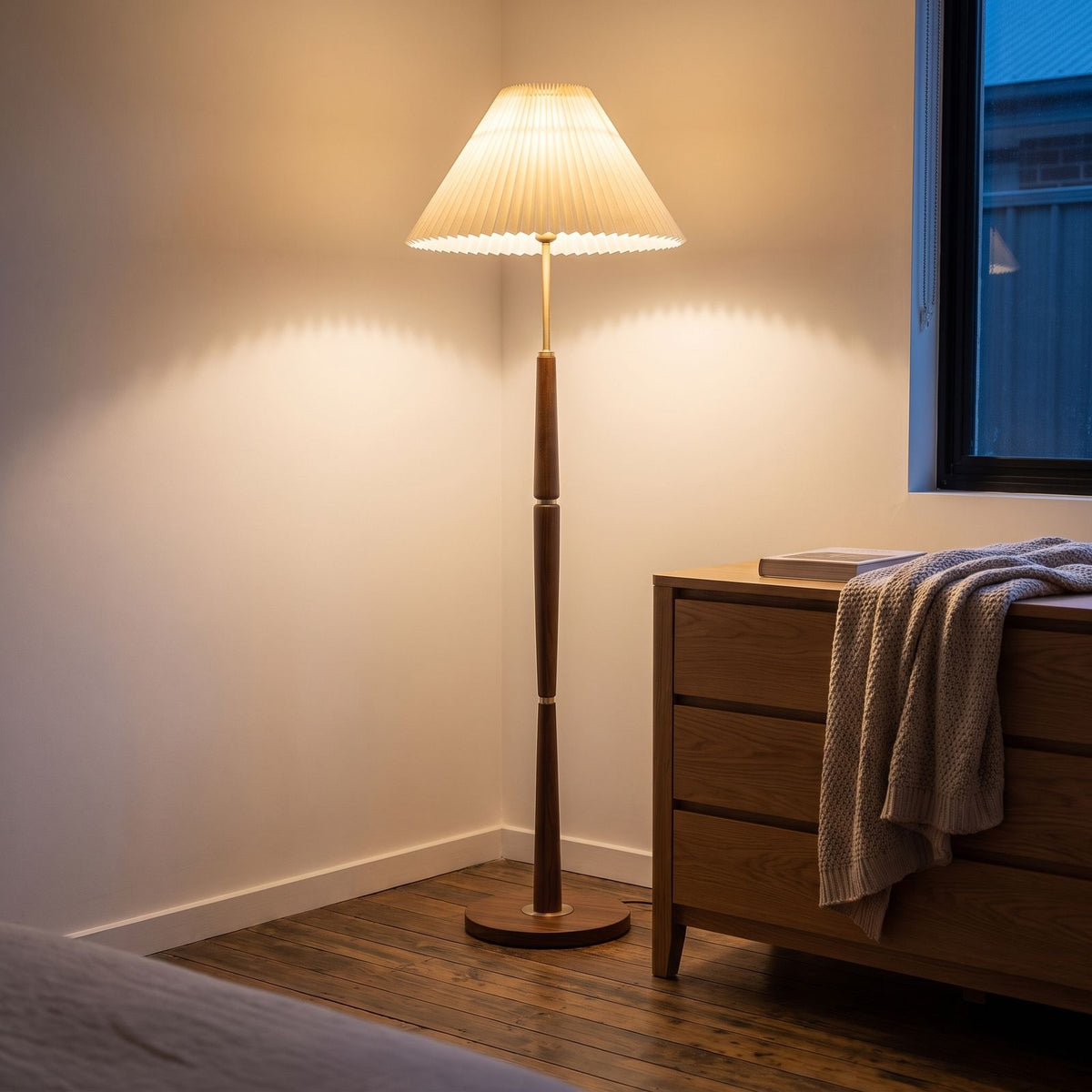 Turned wood floor lamp with pleated shade in a bedroom corner beside a dresser
